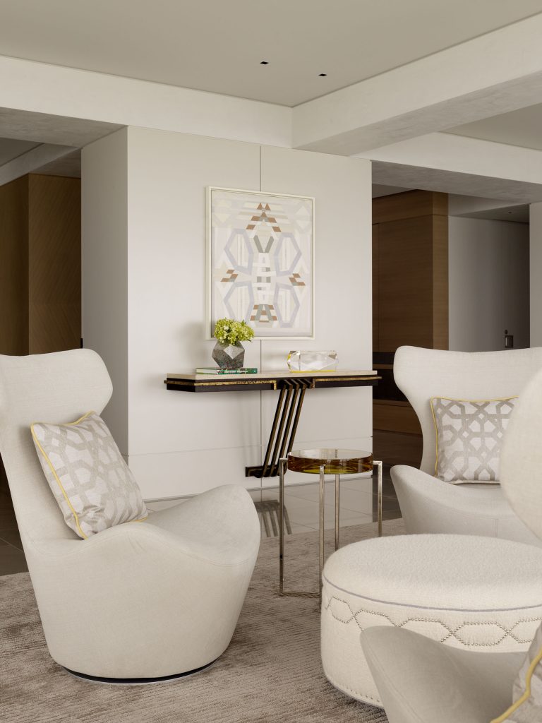 A modern living room with cream swivel chairs, a round ottoman, geometric-patterned pillows, and a soft beige rug. A small side table and a console with decor and flowers sit below abstract framed art on a white wall. The space looks bright and minimal.