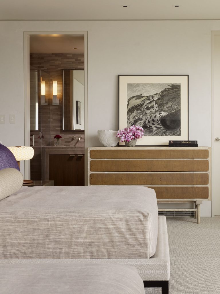 A modern bedroom with beige upholstered furniture, a wooden dresser topped with books, a white bowl, and a vase of pink flowers. Above the dresser hangs a black-and-white abstract artwork. An open door reveals a bathroom with brown tiles and wall-mounted lights.