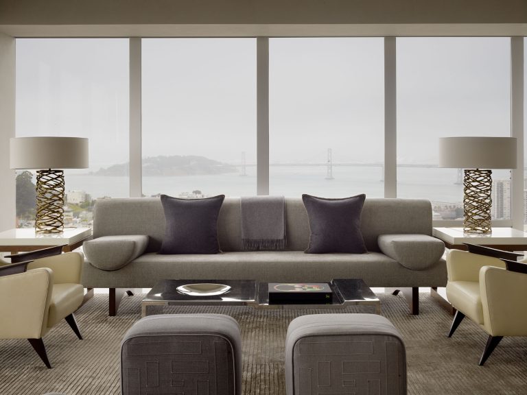 A modern living room with a gray sofa, two dark pillows, geometric ottomans, and a glass coffee table. Two cream armchairs and matching lamps flank the sofa. Large windows reveal a foggy bay and bridge view outside.