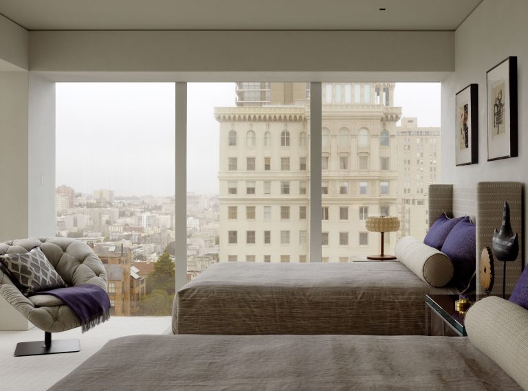 A modern bedroom with two beds dressed in gray linens and purple accent pillows, facing large floor-to-ceiling windows. Outside, tall historic buildings and a cityscape are visible. The room features minimalist decor, framed art, and a cozy, tufted chair.