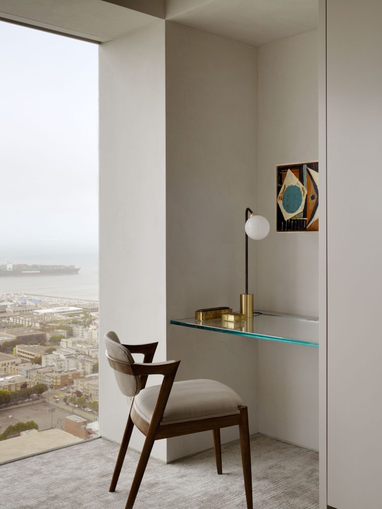 A minimalist home office with a floating glass desk, modern wooden chair with light upholstery, brass lamp, tray, and small abstract wall art. The workspace is by a large window overlooking a cityscape and water in the distance, with soft natural light.