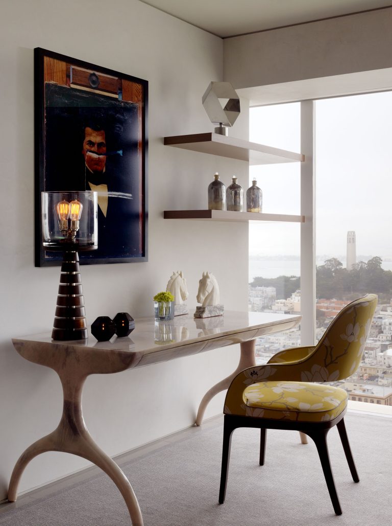 A modern office with a sculptural desk, yellow floral chair, lamp, two black candle holders, and white horse figurines. Art with a suited figure hangs on the wall. Floating shelves hold bottles and a sculpture. Large windows show a cityscape and distant tower.