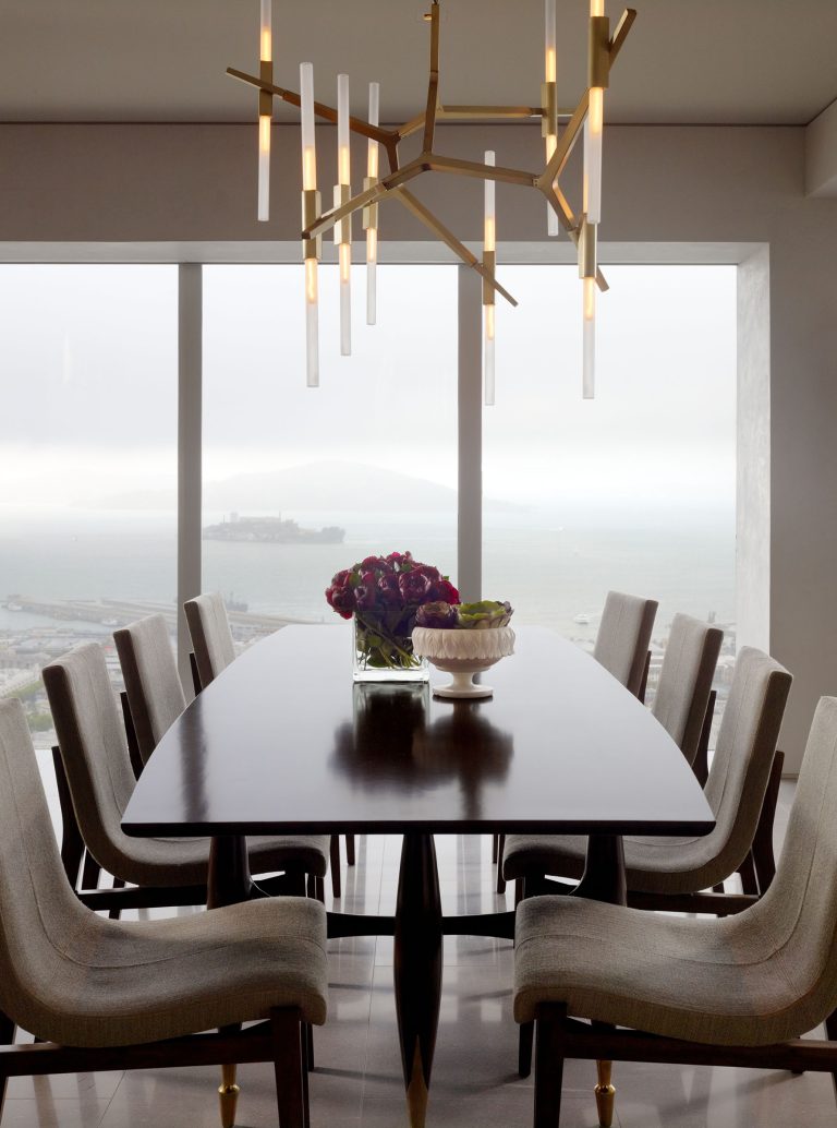 Modern dining room with a long dark wooden table surrounded by eight beige upholstered chairs. A gold geometric chandelier hangs above. The table holds a vase of purple flowers and a cake stand. Large windows reveal a foggy seascape and distant island.