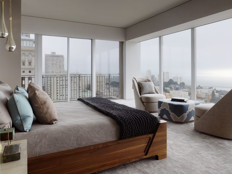 Modern bedroom with a wooden bed, gray bedding, and blue accent pillows. Large floor-to-ceiling windows reveal a cityscape view. A lounge chair and ottoman sit in the corner on a light carpet, creating a serene, minimalist atmosphere.