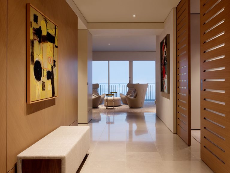 Modern hallway with beige tiled floors, wood-paneled walls, and abstract art. A cushioned bench sits on the left. At the end, two armchairs and a small table face large windows with an ocean view. Wood slat doors are partially open on the right.