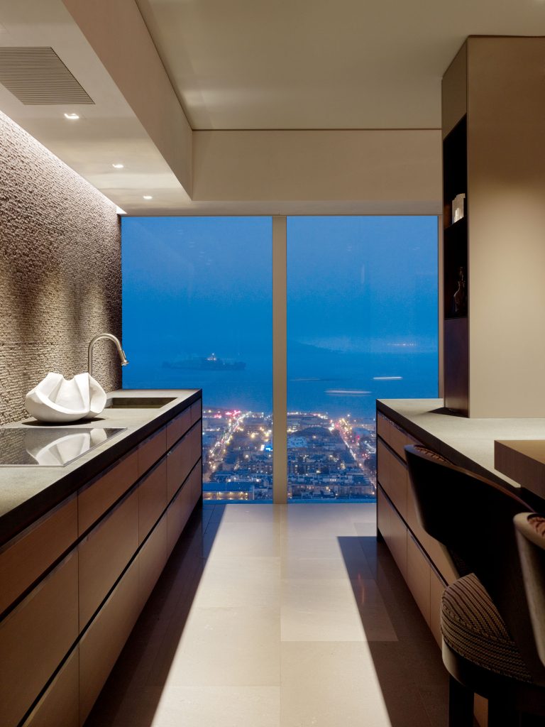 Modern kitchen with sleek dark cabinets and stone countertops, featuring a sculptural white bowl on the counter. Large floor-to-ceiling window offers a cityscape and ocean view at dusk, with illuminated buildings and a calm, blue twilight sky.