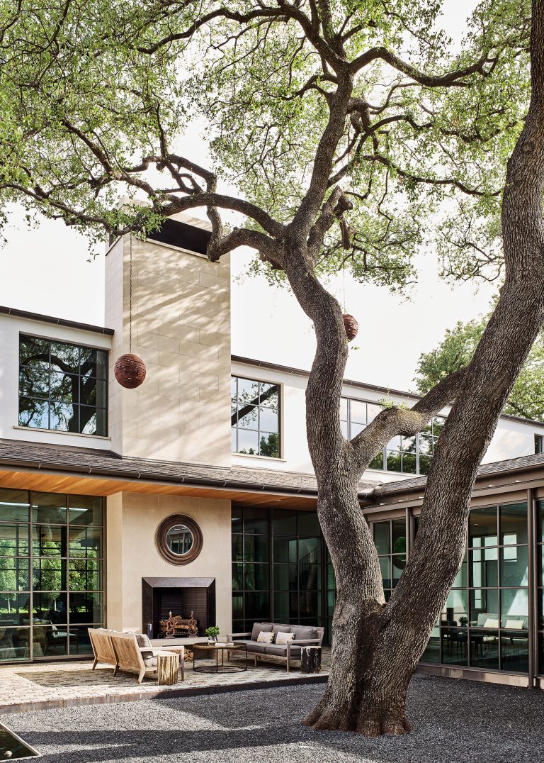 A modern house with large windows and light stone walls surrounds a courtyard. A mature tree with twisting branches stands in the foreground, its base in dark gravel. Outdoor seating and a fireplace are arranged beneath the tree, with two spherical hanging lights.