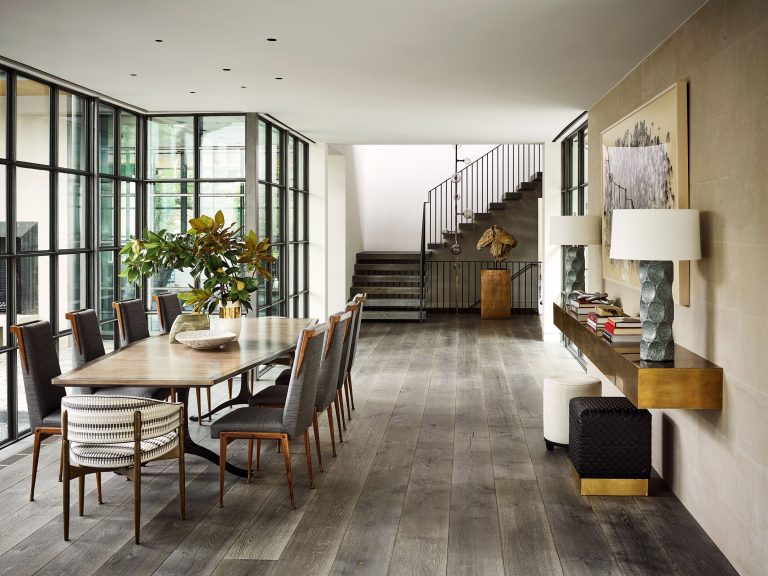 A modern dining room with a wooden table, eight upholstered chairs, two striped armchairs, tall windows on one side, a console table with lamps and decor, wooden floors, a large plant, artwork on the wall, and a staircase in the background.