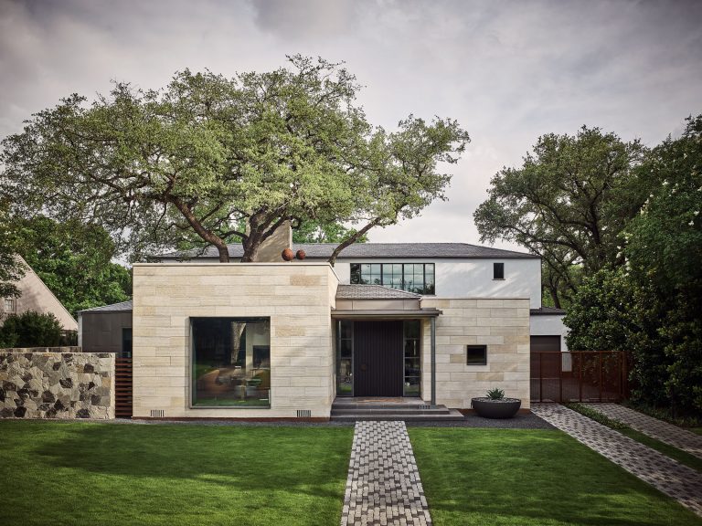 Modern two-story home with light stone facade, large dark-framed windows, and flat roof. A mature tree grows through an opening in the roof. Neatly mowed lawn, stone walkway, low planter, and lush trees surround the house under a cloudy sky.