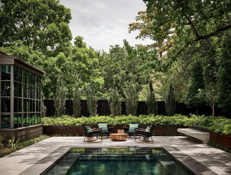 Modern backyard with a small pool in the foreground, four black lounge chairs with striped cushions around a wooden firepit on a concrete patio, lush green plants, tall trees, and a glass-walled house extension on the left. The sky is partly cloudy.