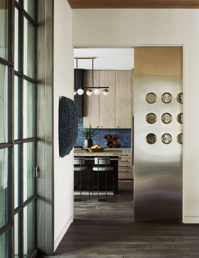 A modern kitchen seen through a doorway features light wood cabinets, a dark blue tile backsplash, and two barstools at a peninsula. A metallic sliding door with round glass inserts and contemporary decor add a stylish touch to the open, airy space.