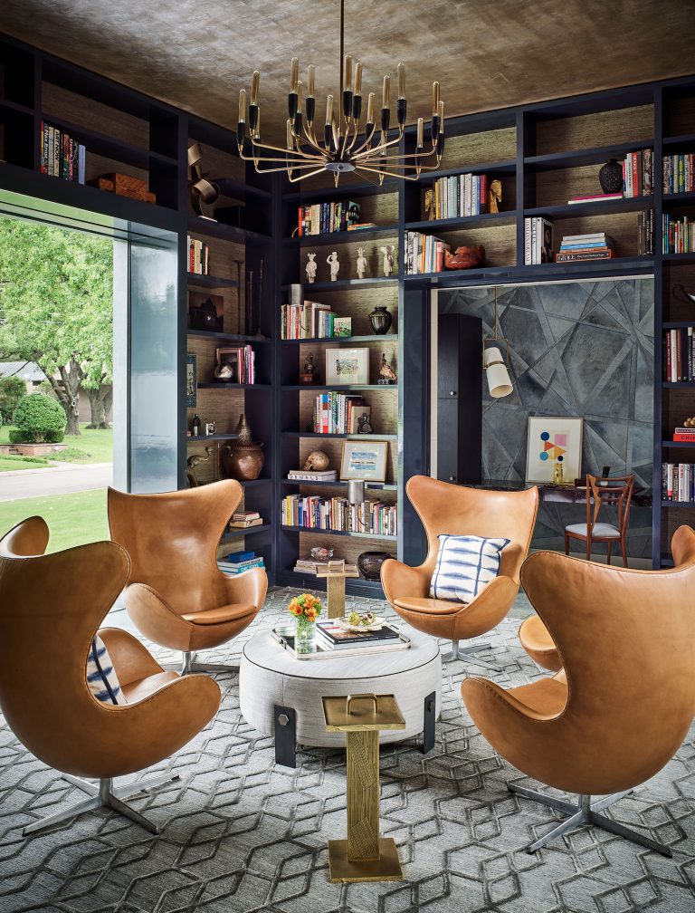 A cozy library with dark built-in bookshelves, tan leather egg chairs arranged in a circle around a round white coffee table, geometric gray rug, modern gold chandelier, and large window showing a green lawn. The room feels bright and inviting.