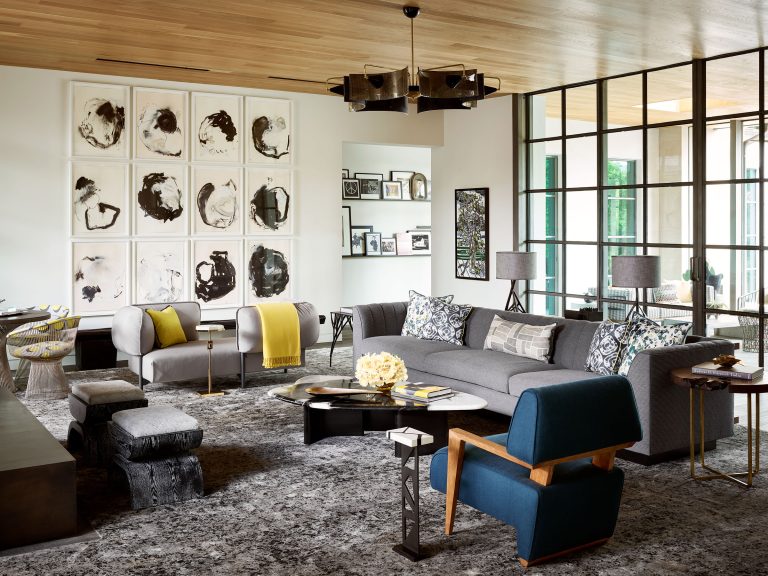 A modern living room with a gray sofa, a blue armchair, and abstract black-and-white art on the wall. The space features a patterned rug, marble coffee table, accent pillows, and throws in yellow and gray tones, and is well-lit by large windows.