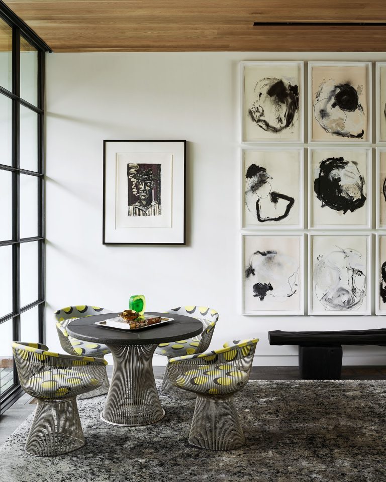 A modern dining nook with three wire chairs with patterned cushions around a round black table. Abstract art hangs on a white wall, featuring black and beige brushstrokes. Large windows, a textured rug, and a wooden ceiling complete the stylish, minimalist space.