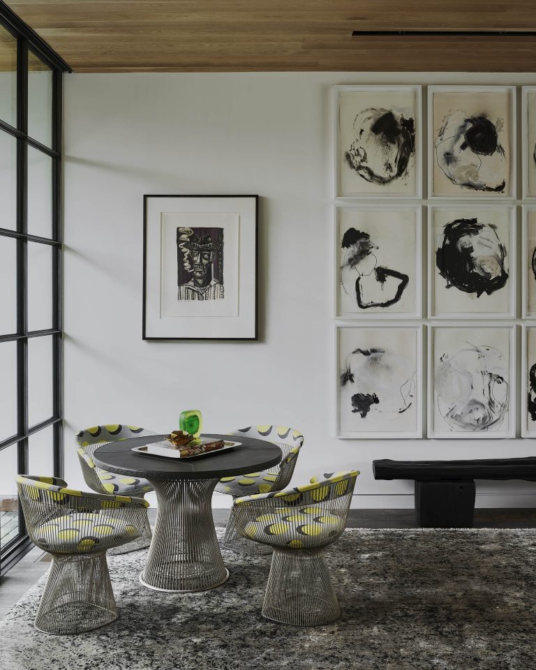 A modern dining nook with a round black table, three wire chairs with gray and yellow cushions, and a large window. Abstract black-and-white art fills one wall, with a framed portrait beside it. A black bench sits below the artwork on a patterned rug.
