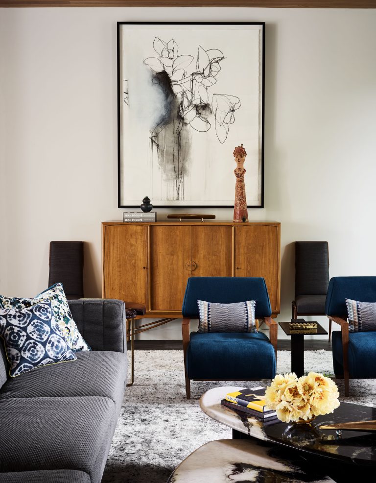 A modern living room with a gray sofa, two blue armchairs, a wooden cabinet, abstract floral art, and decorative objects. The coffee table holds yellow flowers and books. The space features neutral walls, patterned pillows, and a soft gray rug.