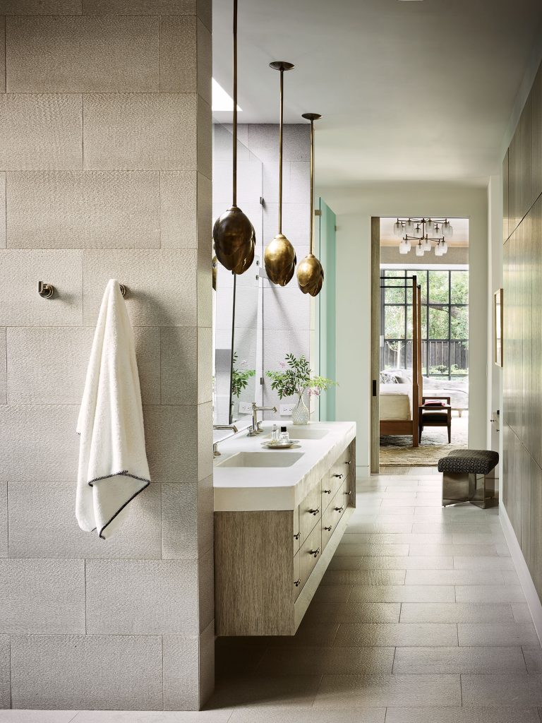 A modern bathroom with beige tiles, a floating double vanity with two sinks, gold pendant lights, and a white towel hanging on a hook. Large windows let in natural light, and a glimpse of a bedroom with a bench and greenery is visible in the background.