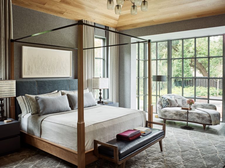 A modern bedroom features a wooden canopy bed with neutral bedding, flanked by two bedside tables with lamps. Large windows offer a view of greenery outside. A cushioned bench sits at the bed’s foot, and a chaise lounge with pillows is near the window.