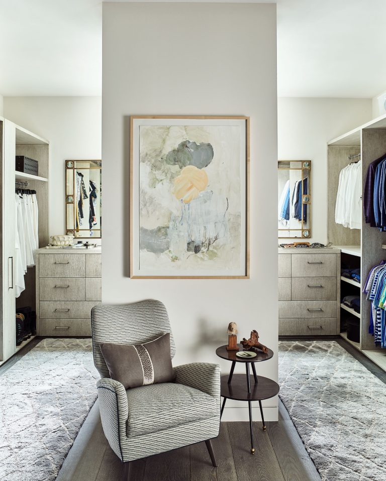 A modern walk-in closet with built-in shelves, hanging clothes, and drawers on both sides. In the center, a gray armchair and round side table sit on a gray rug; above, an abstract art piece in light, neutral colors hangs on a white wall.