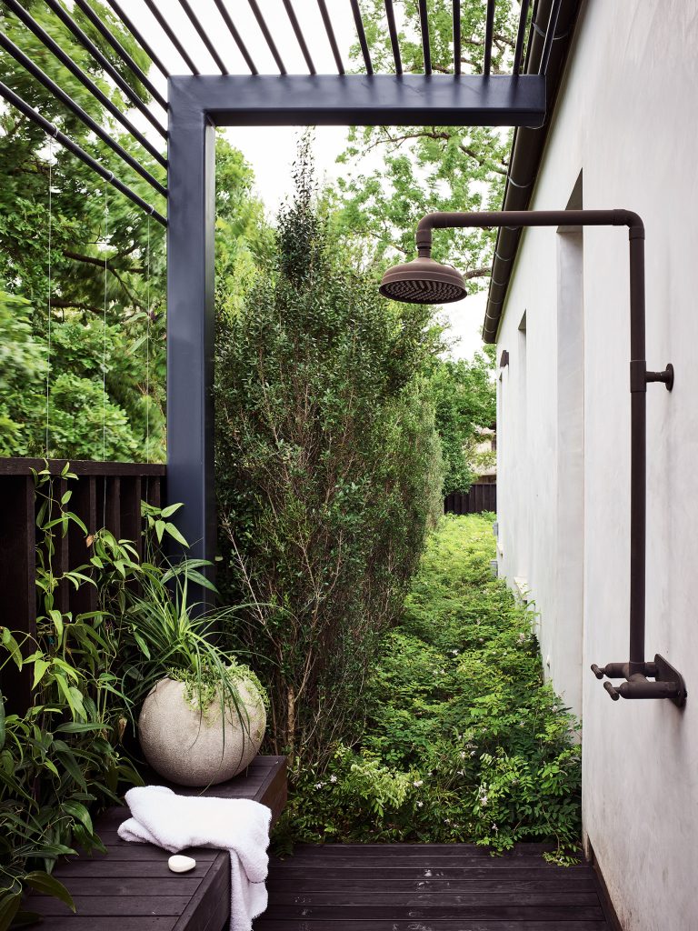 An outdoor shower with a dark metal frame and round showerhead is attached to a light wall. The area is surrounded by lush green plants and trees. A bench holds a round planter with foliage, a white towel, and a bar of soap. Natural light filters through slats above.