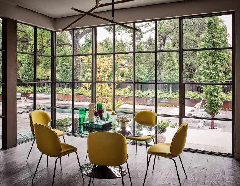 A modern dining area with a round glass table, five yellow chairs, and a geometric chandelier. Floor-to-ceiling windows reveal a lush garden, a pool, and manicured greenery outside, flooding the room with natural light.
