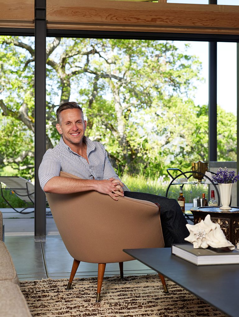 A man with short hair and a striped shirt sits smiling in a modern tan chair in a bright living room with large glass windows. Green trees are visible outside. In front of him is a dark coffee table with books, a shell, and a purple flower arrangement.