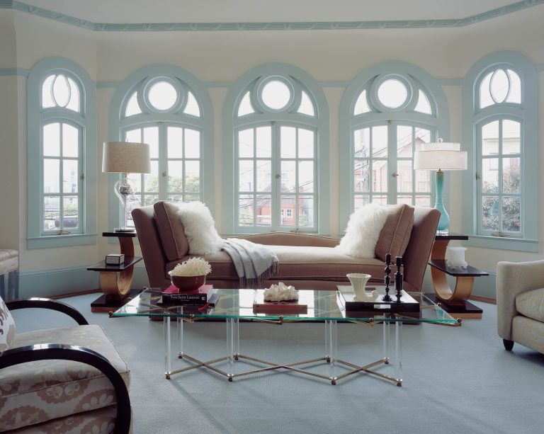 A bright living room with arched windows, a brown chaise lounge with white pillows and throw, glass coffee table with books and decor, two lamps on side tables, and an upholstered chair, all in soft pastel tones and abundant natural light.