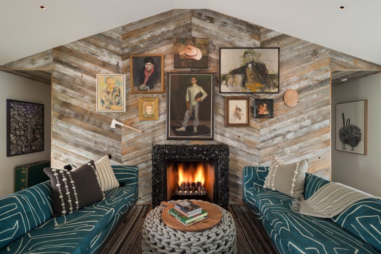 A cozy living room with two patterned teal sofas facing each other, a round knit ottoman coffee table, and a rustic wood-paneled wall. A black ornate fireplace with a fire burning sits below an eclectic gallery of portraits and artwork. Pillows add warmth.