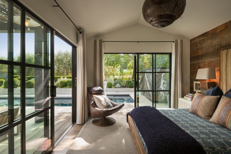 A cozy bedroom with a modern, earthy design features a large bed, a brown chair, and glass sliding doors opening to a patio and pool. Lush greenery and trees are visible outside; natural light fills the room, creating a tranquil, airy atmosphere.