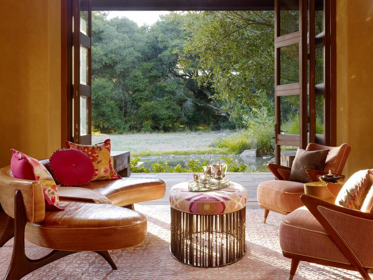 Two modern lounge chairs with colorful cushions face a round ottoman topped with a silver tea set. Large open doors reveal a sunlit garden with lush green trees and grass, creating a cozy, inviting indoor-outdoor living space.