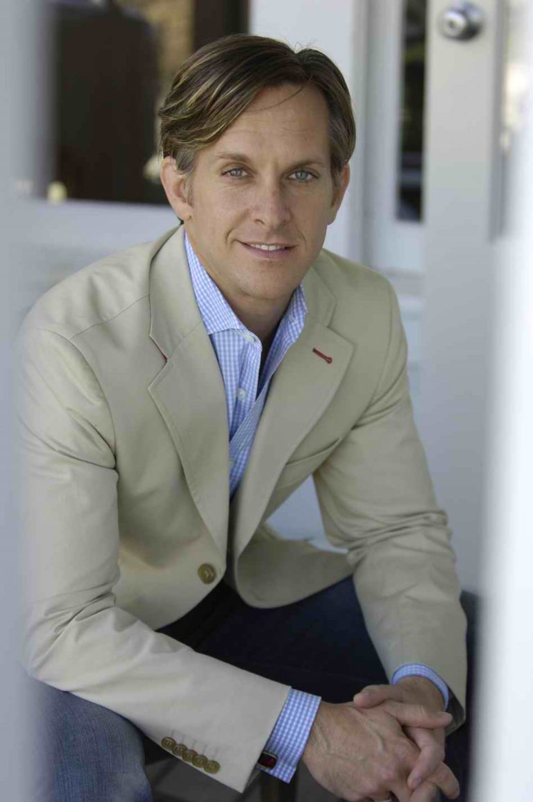 A man with light brown hair, wearing a beige blazer over a blue checkered shirt, sits with hands clasped. He is looking at the camera and smiling softly. The background is out of focus, showing a white door and wall.