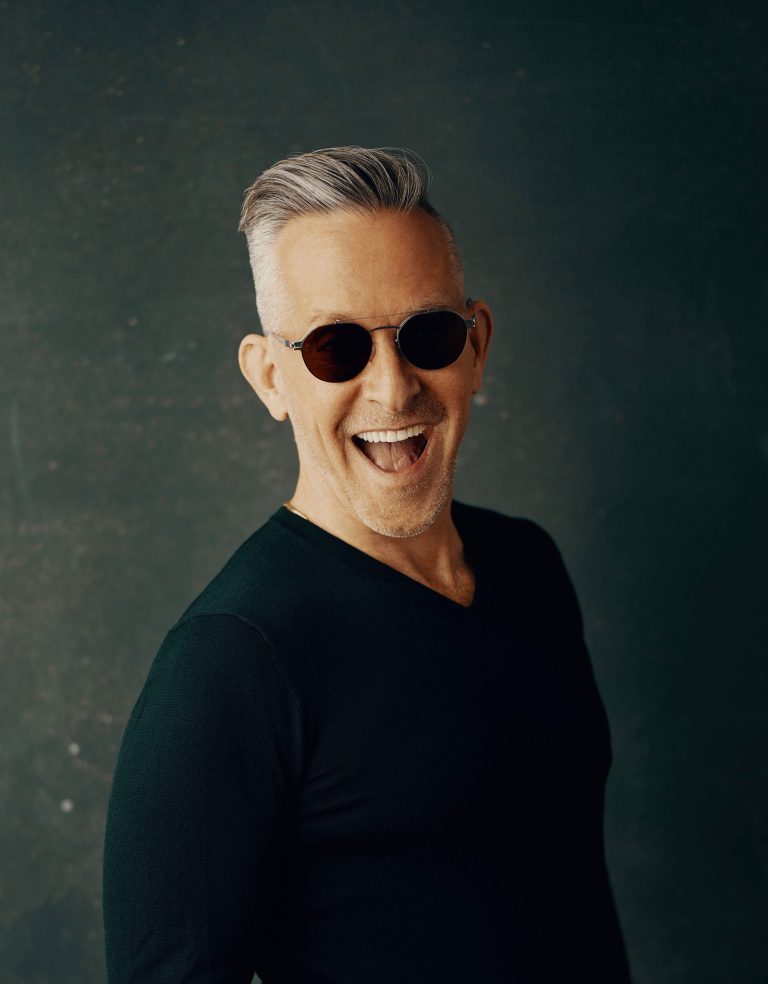 A smiling older man with gray hair styled back and short on the sides wears round black sunglasses and a fitted black shirt, standing against a dark textured background. He faces slightly left and has a playful, open-mouthed expression.