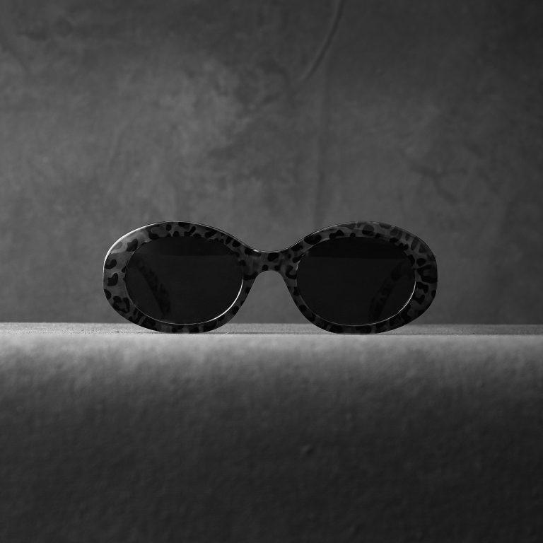 Black and white photo of oval-shaped sunglasses with dark lenses and a leopard print frame, centered on a textured surface against a softly lit, textured background. The image has a minimalist and moody aesthetic.