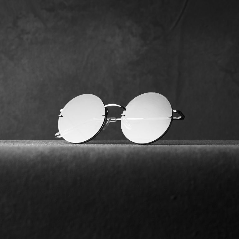 A pair of round, reflective glasses with thin metal frames sits on a textured surface. The image is in black and white, with soft lighting casting gentle shadows and highlighting the minimalist, vintage style of the glasses against a plain, dark background.