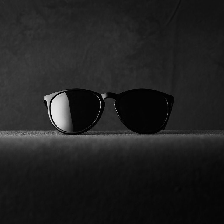 A pair of black sunglasses is centered on a dark, textured surface with a plain, dark background. The lighting casts a faint reflection on the left lens, highlighting the sleek and minimalist design of the sunglasses.