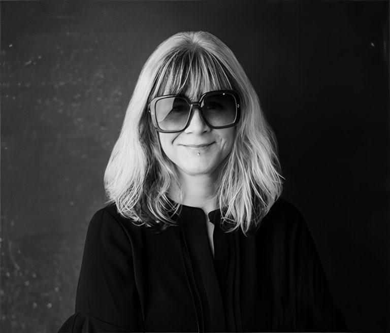Black and white photo of a woman with shoulder-length, light-colored hair, wearing large dark sunglasses and a dark blouse. She is smiling softly and standing against a plain, slightly textured background.