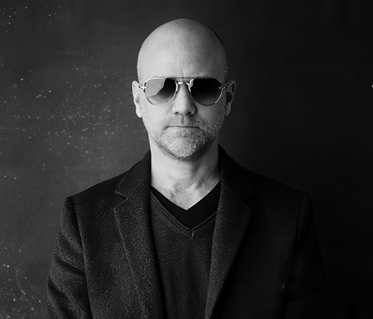 A middle-aged man with a bald head and light facial stubble wears aviator sunglasses, a dark blazer, and a V-neck shirt. He stands against a dark, textured background, facing forward with a neutral expression. The photo is in black and white.
