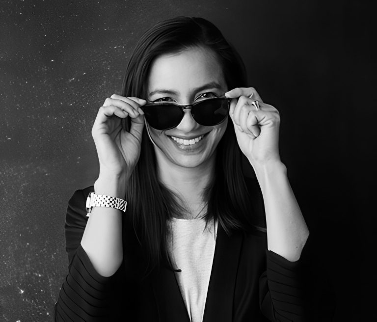 A smiling woman with straight dark hair holds sunglasses just below her eyes. She wears a light shirt, a dark blazer, and a checkered wristwatch. The background is dark with subtle speckles, and she looks directly at the camera.