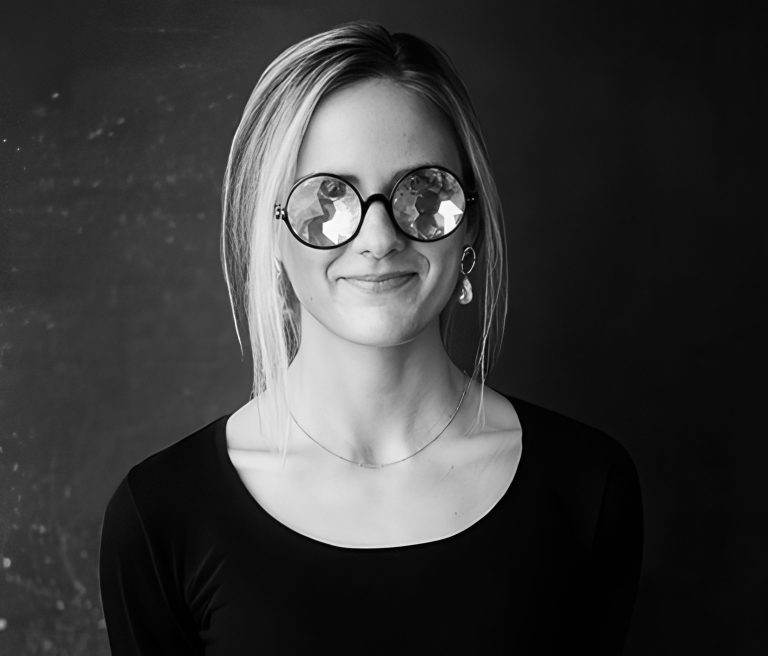 A woman with straight, light hair wears large, round, reflective glasses and a black top. She has a slight smile, a single dangling earring, and stands against a dark, textured background. The photo is black and white.
