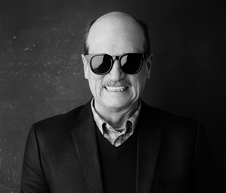 A middle-aged man with a bald head, mustache, and wearing dark sunglasses smiles at the camera. He is dressed in a dark blazer, sweater, and collared shirt. The background is plain and dark, highlighting his figure.